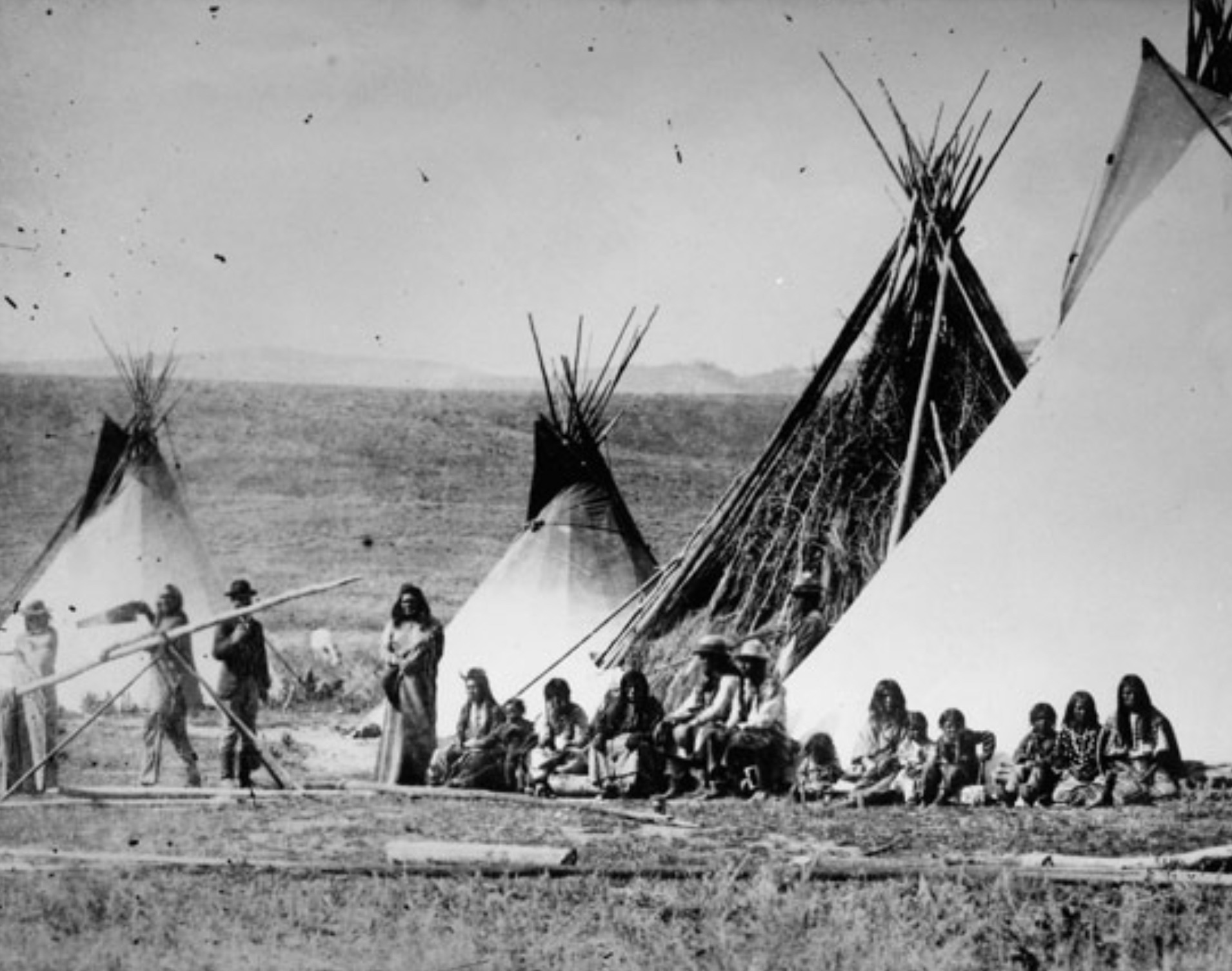 Native American men and women of the Shoshone tribe pose near tepees in Wyoming Territory. Chief Washakie stands with a wooden shaft and a saw. 